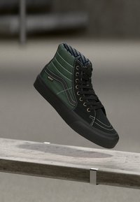 Schwarze und dunkelgrüne High-Top-Sneaker mit weißen Nähten und schwarzen Schnürsenkeln, die über einer Betonbank schweben.