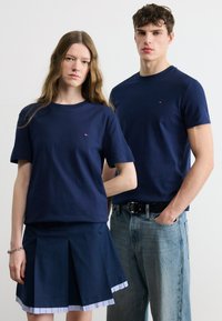 Νεαρή γυναίκα και άνδρας που φορούν μπλε μπλουζάκια με στρογγυλή λαιμόκοψη. Η γυναίκα φοράει πλισέ φούστα με βραχιόλι, ο άνδρας ανοιχτό μπλε τζιν με το χέρι στην τσέπη.