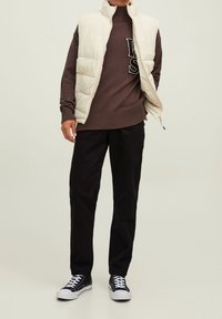 Kamizelka puchowa w kolorze kremowym, brązowy sweter w prążki z logo, czarne spodnie i czarne sneakersy z białymi podeszwami. Prosty, warstwowy wygląd.