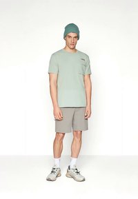 Lys grønn T-skjorte med korte ermer og brystlomme, kombinert med lysegrå shorts. Modellen har på seg en petroleumfarget strikkelue og grå joggesko.