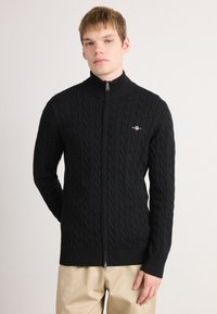 Fekete, kötött, zipzáros pulóver kábelmintával, magas gallérral és bordázott mandzsettákkal és aljjal. A bal oldalon egy kis logó található.