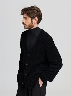 Cardigan nero e peloso con scollo a V profondo, maniche lunghe e chiusura con quattro bottoni. Indossato sopra un dolcevita nero, con vestibilità rilassata.