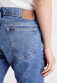 Närbild av en persons hand nära bakfickan på blå Lee jeans med synliga märkesetiketter och sömnadsdetaljer.