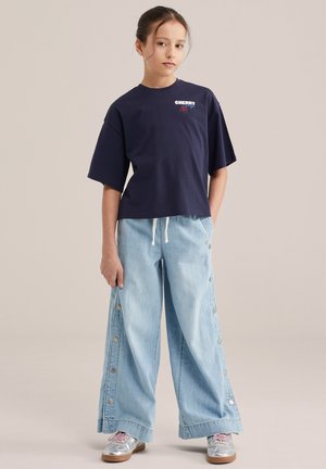 Mlajša oseba stoji oblečena v mornarsko modro majico s tekstom "CHERRY", široke svetlo modre kavbojke s stranskimi gumbi in kovinsko srebrne superge.