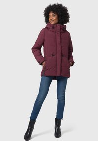 Burgundy puffermantel med justerbar huva och dragsko, med två sidfickor och kviltat mönster. Kombineras med blå jeans och svarta stövlar.