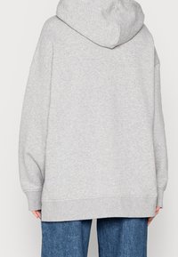 Eine Person trägt einen lockeren, hellgrauen Kapuzenpullover und eine blaue Jeans, von hinten vor einem schlichten Hintergrund gezeigt.