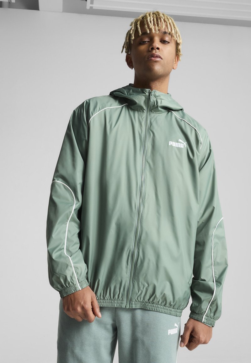 Grüne, leichte Jacke mit weißen Paspeln, Kordelzugkapuze und elastischen Bündchen. Verfügt über einen Reißverschluss und ein Logo auf der Brust.