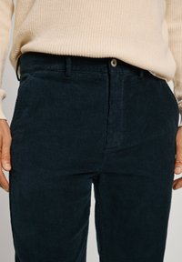Person wearing navy blue corduroy pants and a beige knitted sweater, hands relaxed by their sides.