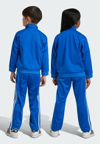 Blå sportiga träningsoveraller med hög krage, ribbade ärmmuddar och tre vita ränder längs sidorna, uppbackade med vita och svarta sneakers.