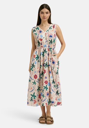 Frau trägt ein ärmelloses beiges Midikleid mit bunten Blumen- und Sonnenmustern, elastischer Taille und Sandalen, steht mit einer Hand in der Tasche.