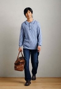 Pull en laine bleu clair avec un motif en chevrons, associé à un jean foncé et un sac de voyage en cuir marron. Des chaussures sombres complètent l'ensemble.