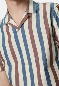 Camicia a maniche corte con colletto, caratterizzata da righe verticali in rosso, blu e crema. Tessuto testurizzato con chiusura a bottoni e una vestibilità rilassata.
