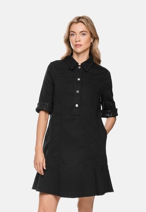 Blonde woman wearing a black button-up dress with rolled sleeves and a flared hem standing against a plain light background.