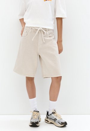 Person iført beige knælange shorts med snøre i taljen, hvide sokker og sorte og hvide løbesko.