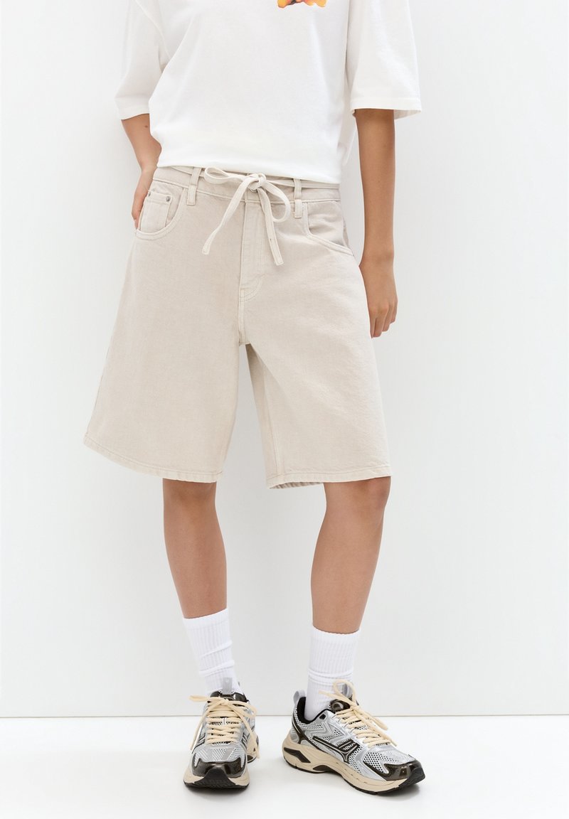 Personne portant un short beige longueur genou avec une taille à cordon, des chaussettes blanches et des chaussures de course noires et blanches.