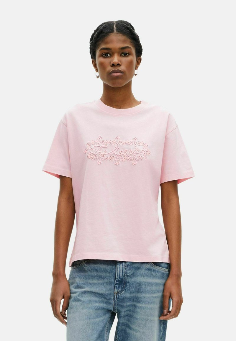 Camiseta de algodón rosa con mangas cortas, que presenta un diseño de texto en relieve en el centro. Combinada con jeans ligeros de mezclilla desgastados.