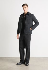 Schwarze gepolsterte Jacke mit einem Frontreißverschluss, zwei Brusttaschen und einem Kragen, kombiniert mit schwarzen weit geschnittenen Hosen und schwarzen Schuhen vor einem hellen Hintergrund.