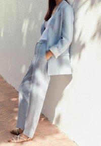 Costume sur mesure bleu clair en lin, composé d'un long blazer et d'un pantalon droit. Les sandales présentent un design à lanières avec des accents subtils.