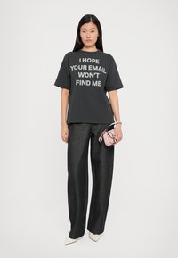Grå bomulds t-shirt med hvid tekst, parret med sorte brede bukser. Modellen holder en lille lyserød håndtaske og har cremefarvede spidse sko på.