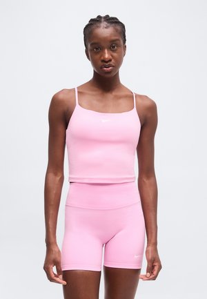 Mladá žena s vrkočmi v svetloružovom Nike crop tope a zodpovedajúcich vysokých cyklistických šortkách na jednofarebnom pozadí.