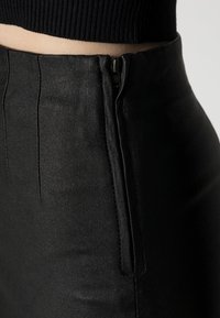 Pantalon noir avec une texture lisse et légèrement brillante, présentant une fermeture éclair et des détails de coutures verticales à la taille.
