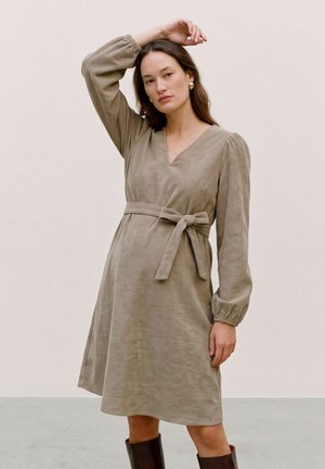 Une femme pose devant un fond de studio uni, vêtue d’une robe taupe ceinturée à manches longues, de boucles d’oreilles dorées et de bottes foncées montant jusqu’aux genoux.