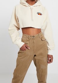 Osoba má na sobě krémovou krátkou mikinu s logem "ellesse" a světle hnědé kalhoty cargo s vysokým pasem a detaily záplat.