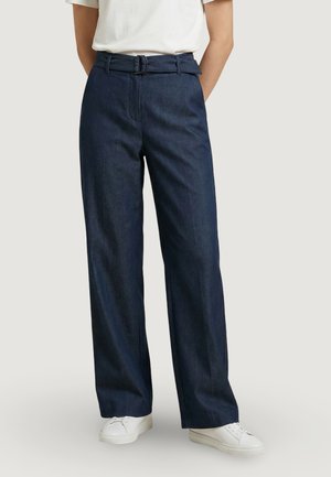 Persona che indossa pantaloni a gamba larga a vita alta di colore blu scuro con cintura e sneaker bianche, in piedi contro uno sfondo chiaro uniforme.