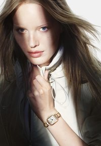 Junge Frau mit langen Haaren, die den Kragen einer weißen Jacke festhält, trägt eine goldene Armbanduhr mit quadratischem Ziffernblatt und blickt mit Licht auf ihr Gesicht nach vorne.