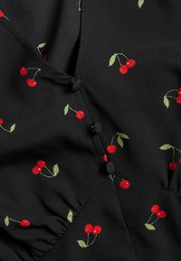 Zwarte stoffen top met een rood kersenpatroon, groene bladeren en delicate knoopdetails. Heeft een geraffineerde structuur langs de naden.