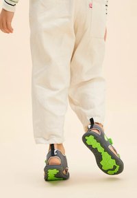 Sandali aperti grigi con accenti verdi, suole testurizzate e cinturini regolabili, indossati con pantaloni chiari e a sigaretta.