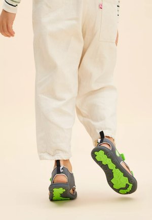 Graue Sandalen mit offenen Zehen und grünen Akzenten, mit strukturierten Sohlen und verstellbaren Riemen, getragen zu hellen, schmal zulaufenden Hosen.