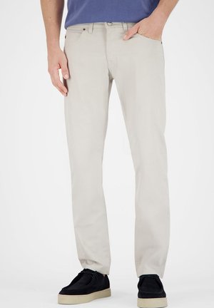 Helle beige Hosen mit geradem Schnitt, aus Baumwollstoff hergestellt. Sie verfügen über frontale Taschen und einen klassischen Knopf- und Reißverschluss. Getragen mit schwarzen Schuhen.