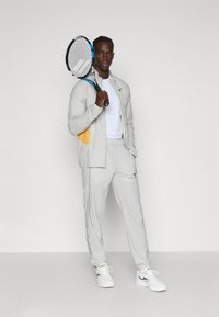 Lichtgrijze sportieve trainingsoutfit met een gele accent, gedragen met een wit shirt. Het onderwerp houdt een tennisracket vast en draagt witte sneakers.