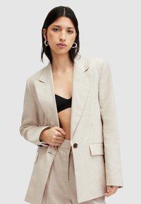 Blazer in lino beige chiaro con rever a lancia, chiusura con un bottone e tasche laterali. Indossato sopra un top nero, mette in evidenza il suo fit rilassato.