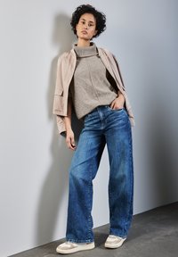 Giacca in similpelle beige, maglione a collo alto in maglia con motivo testurizzato e jeans azzurri a gamba larga. Sneakers beige casual.