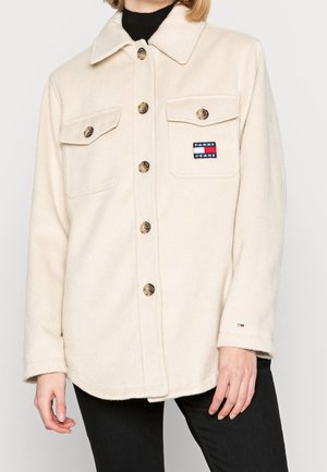 Frau trägt eine cremefarbene Jacke mit Knöpfen und Brusttaschen mit Tommy Jeans Logo, kombiniert mit schwarzen Hosen und einem schwarzen Rollkragen.
