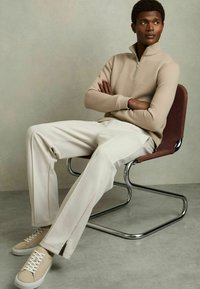 Beige svetr na zip s ribovým vzorem, krémové široké kalhoty a béžové tenisky, sedící na moderní chromové židli s burgundským sedákem. Hladký pozadí.