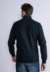 Czarna bluza z teksturowymi panelami na ramionach, długimi rękawami i ściągaczem u dołu. Noszona z niebieskimi dżinsami, przedstawiająca widok z tyłu.