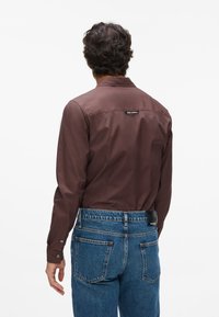 Camicia marrone a maniche lunghe con colletto button-down, caratterizzata da un'etichetta logo sul retro, abbinata a jeans in denim blu con dettagli delle tasche posteriori.