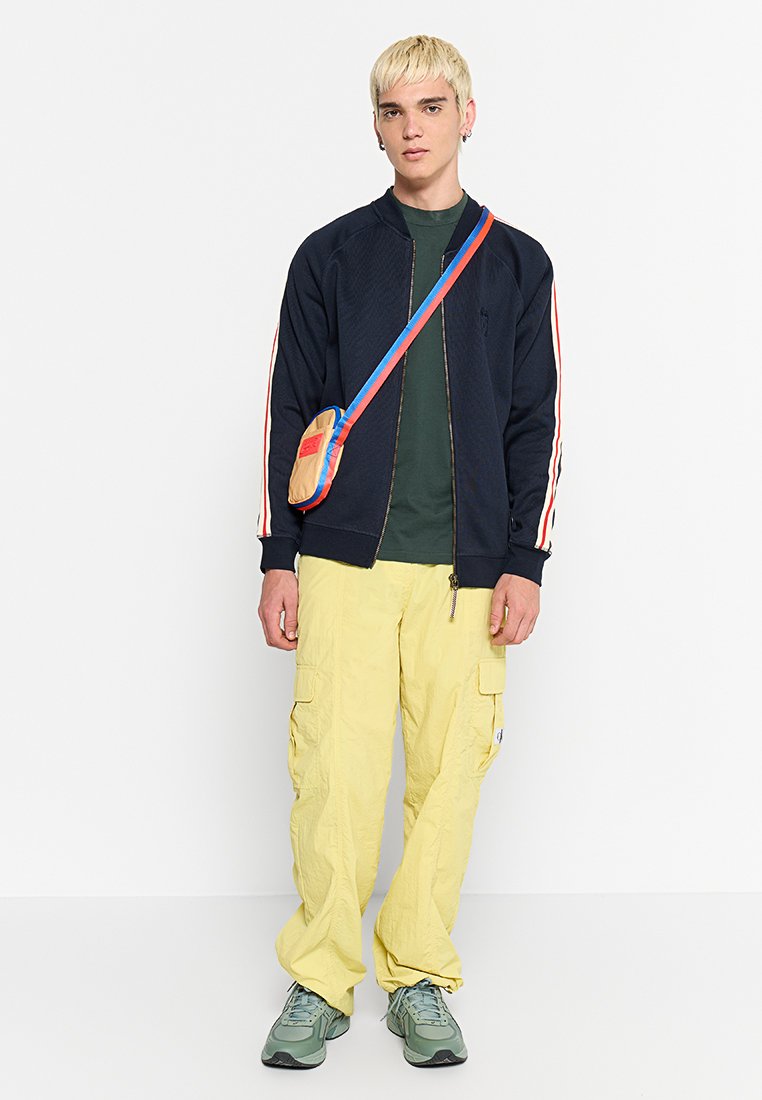 Marineblaues Zip-Up-Jackett mit roten und weißen Streifenakzenten, grünes T-Shirt, gelbe Cargo-Hosen und eine kleine, mehrfarbige Umhängetasche.