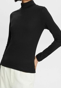 Person som bär en åtsittande svart långärmad polotröja och ljusa byxor, med den ena handen vilande på höften.