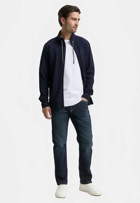 Veste zippée bleu marine avec deux poches avant, portée sur un T-shirt blanc, associée à un jean foncé et des baskets blanches. La veste possède un col montant.