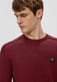 Bordeauxroter gestrickter Pullover mit Rundhalsausschnitt, gerippten Bündchen und einem schwarzen Logo-Patch auf der Brust. Glatte Textur, taillierter Schnitt, ohne Muster.