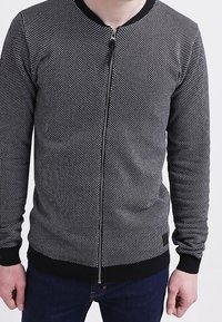 Svart och vit prickig zip-up jacka med ribbade mudd och fåll, med rund halsringning och en slimmad, åtsittande design.