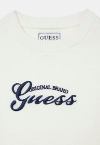 Pull en maille blanche avec col côtelé et logo "Guess" brodé en bleu marine, surmonté du texte "Original Brand".