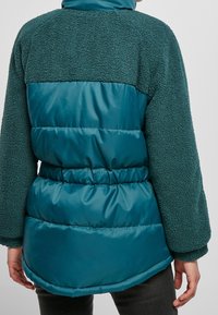 Tyrkysová péřová bunda s huňatým strukturovaným materiálem na ramenech a rukávech, hladká spodní část a elastický pas pro lepší padnutí.