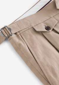 Beige byxor med veck, med en metallspänne och brun knapp. Tyget har en jämn struktur, med en kontrasterande innerfoder.