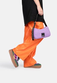 Borsa a tracolla in finta pelle viola con un'etichetta nera. Pantaloni lucidi arancioni brillanti, sneaker colorate con un motivo e suola spessa.