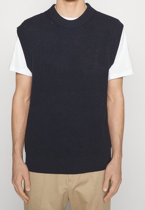 Homme portant un gilet en tricot noir sans manches sur un t-shirt blanc et un pantalon beige, debout devant un fond neutre.